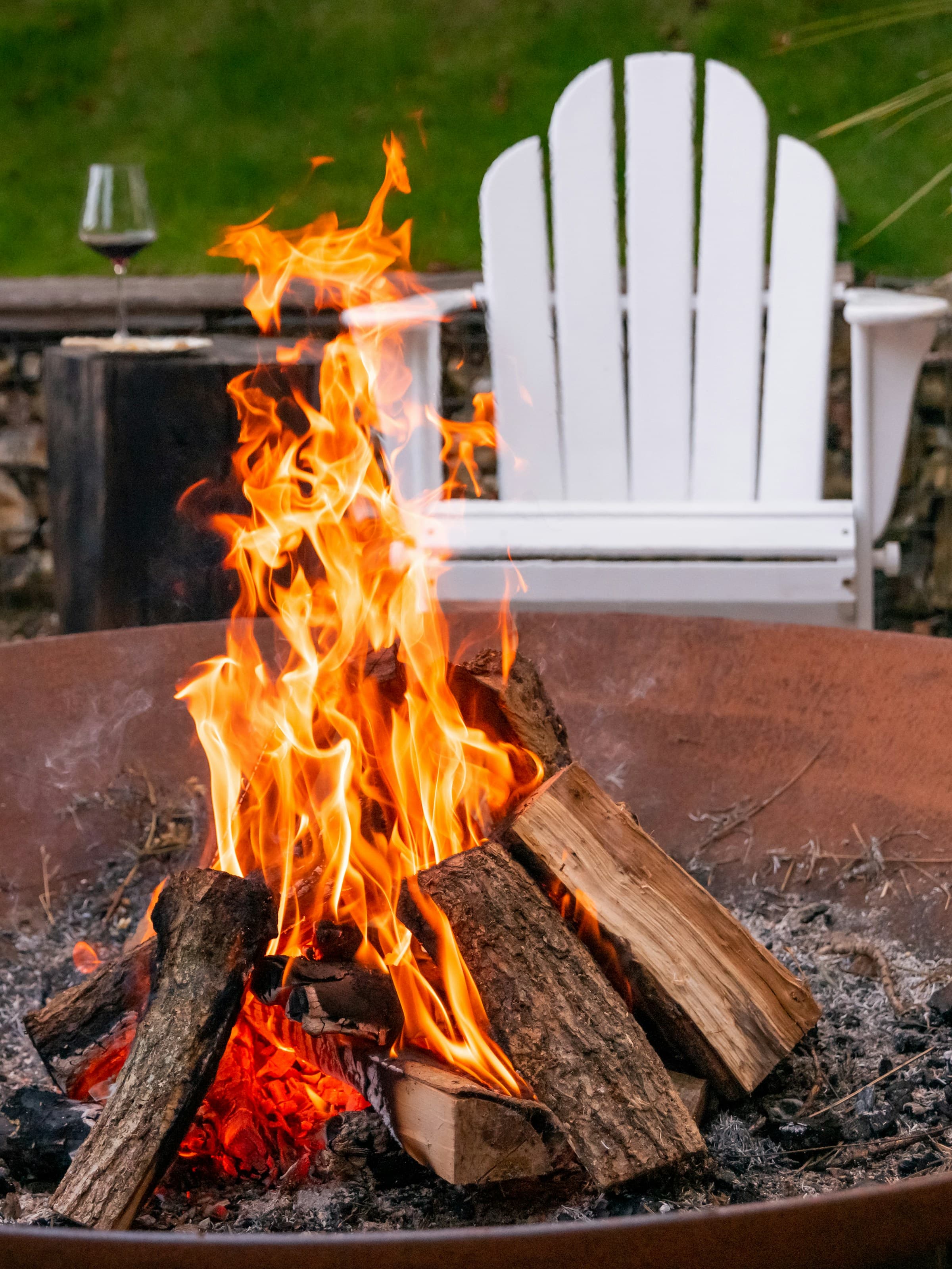Brennendes Holz in einer Feuerschale, dahinter ein Glas Wein auf einer Steinmauer
