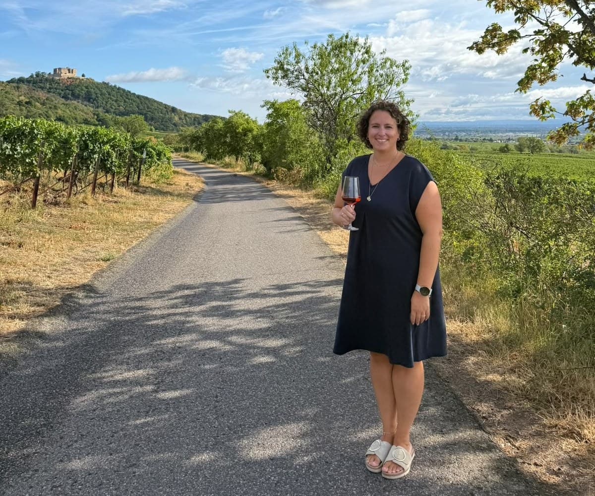 Gastgeberin von PfalzErlebnisse mit einem Glas Wein auf einem Weinbergweg, im Hintergrund das Hambacher Schloss